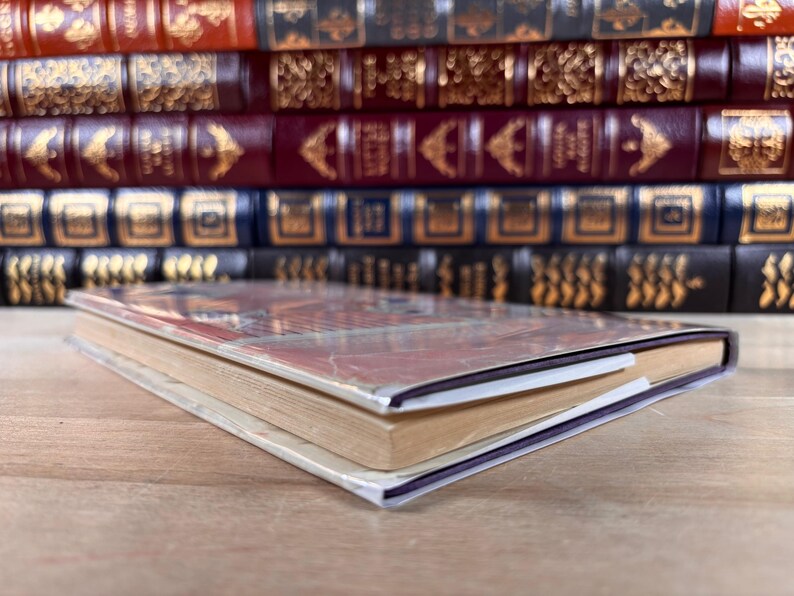 May include: A single paperback book with a reddish-brown cover lies flat in the foreground. Behind it, a row of five hardcover books with ornate, gold-embossed spines in shades of red, burgundy, and dark blue are arranged horizontally on a shelf.