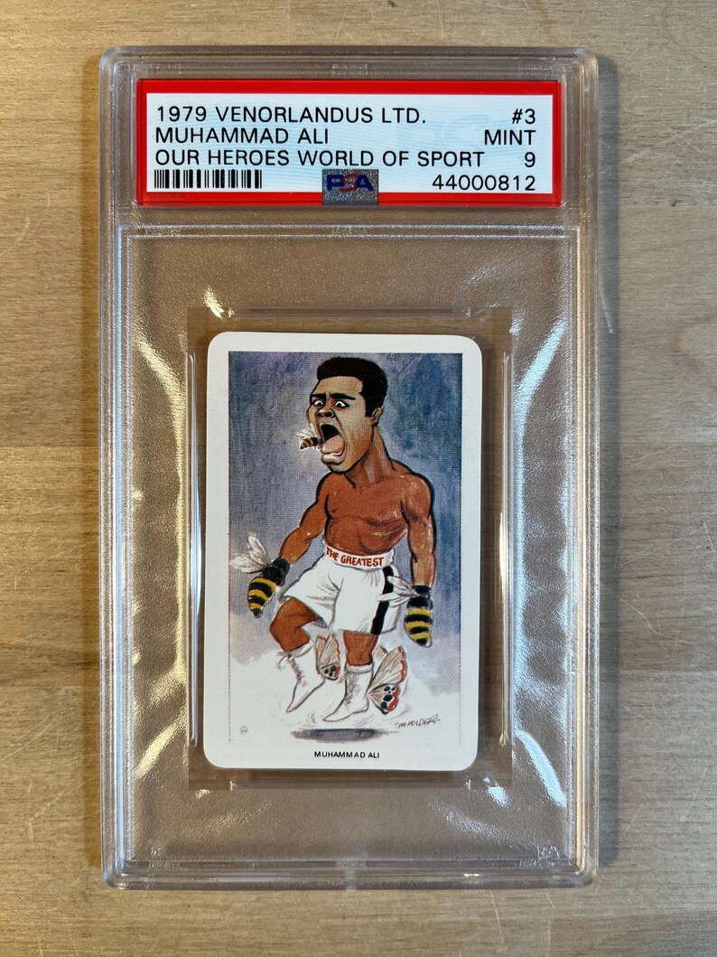 May include: A vintage trading card featuring a cartoon illustration of Muhammad Ali, a famous boxer, with the text "The Greatest" on his boxing shorts. The card is from the 1979 Venorlandus Ltd. "Our Heroes World of Sport" series.
