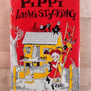 May include: A hardcover book titled Pippi Longstocking by Astrid Lindgren with a bright red cover. The illustration features a yellow house, a spotted horse on a porch, a monkey on a ladder, and a girl with red braids in a red dress.