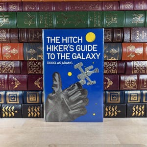 Könnte beinhalten: Ein Buch mit dem Titel "The Hitchhiker's Guide to the Galaxy" von Douglas Adams. Das Buch hat einen blauen Einband mit einem gelben Kreis und einer silbernen Raumschiff-Illustration. Es steht vor einem Stapel alter Bücher.