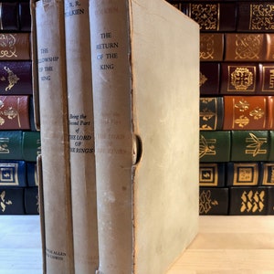 May include: A three-volume set of "The Lord of the Rings" by J.R.R. Tolkien, in a slipcase. The books are bound in brown paper with gold lettering. The slipcase is white with gold lettering.
