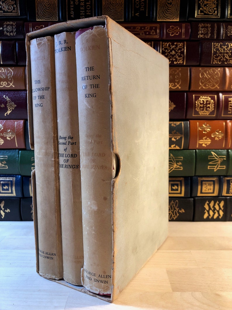 May include: A three-volume set of "The Lord of the Rings" by J.R.R. Tolkien, in a slipcase. The books are bound in brown paper with gold lettering. The slipcase is white with gold lettering.