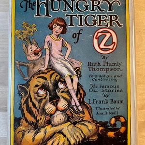 K&ouml;nnte beinhalten: Der Einband eines Vintage-Buches mit dem Titel "The Hungry Tiger of Oz" von Ruth Plumly Thompson. Der Einband zeigt einen Comic-Tiger, auf dessen R&uuml;cken ein M&auml;dchen sitzt. Der Tiger sieht traurig aus und das M&auml;dchen schaut den Betrachter an. Das Buch ist illustriert von Jno. R. Neill.