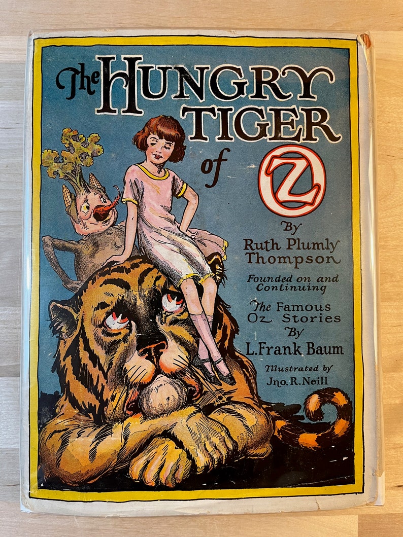 K&ouml;nnte beinhalten: Der Einband eines Vintage-Buches mit dem Titel "The Hungry Tiger of Oz" von Ruth Plumly Thompson. Der Einband zeigt einen Comic-Tiger, auf dessen R&uuml;cken ein M&auml;dchen sitzt. Der Tiger sieht traurig aus und das M&auml;dchen schaut den Betrachter an. Das Buch ist illustriert von Jno. R. Neill.