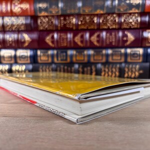 May include: A yellow-covered book lying flat on a surface, positioned in front of a stack of multiple hardcover books with dark red, blue, and black spines featuring gold-colored decorative patterns and lettering on their spines.