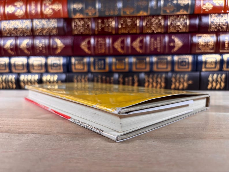 May include: A yellow-covered book lying flat on a surface, positioned in front of a stack of multiple hardcover books with dark red, blue, and black spines featuring gold-colored decorative patterns and lettering on their spines.