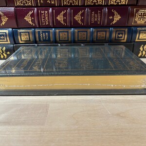 May include: A dark green hardcover book with gold lettering on the spine. The book is titled "Little Women" by Louisa May Alcott. The book is sitting on a wooden surface.