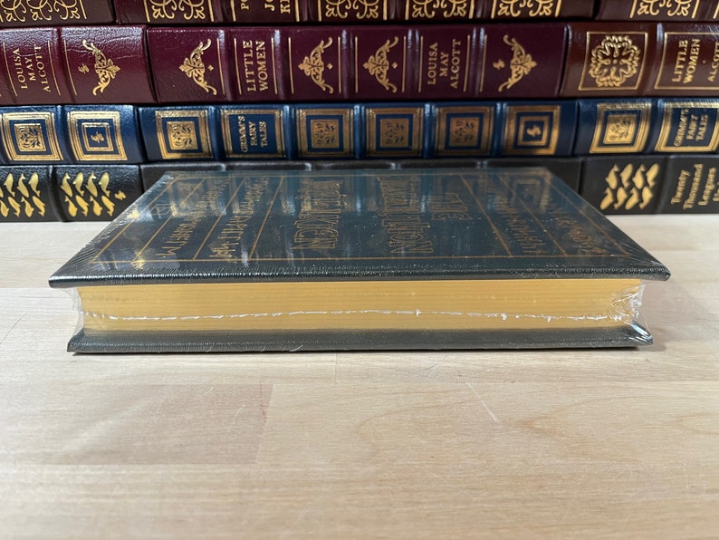 May include: A dark green hardcover book with gold lettering on the spine. The book is titled "Little Women" by Louisa May Alcott. The book is sitting on a wooden surface.