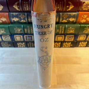 K&ouml;nnte beinhalten: Der R&uuml;cken eines antiken gebundenen Buches mit dem Titel "The Hungry Tiger of Oz" mit einer blauen Illustration eines Tigerkopfes auf wei&szlig;em Hintergrund.