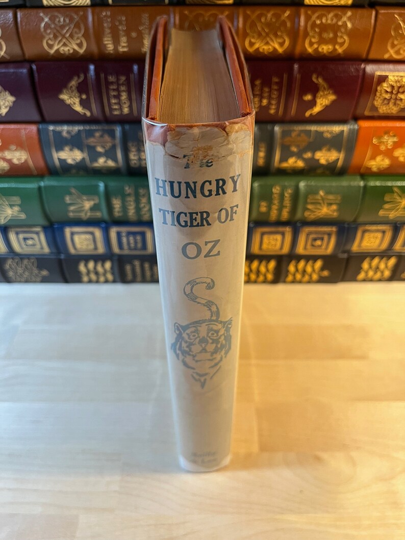 K&ouml;nnte beinhalten: Der R&uuml;cken eines antiken gebundenen Buches mit dem Titel "The Hungry Tiger of Oz" mit einer blauen Illustration eines Tigerkopfes auf wei&szlig;em Hintergrund.