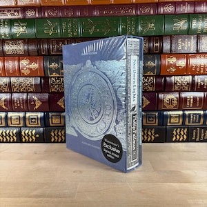 Peut inclure: Un livre bleu intitulé "Northern Lights" de Philip Pullman, une édition limitée signée exclusive de Waterstone's. Le livre présente un motif argenté en relief et est enveloppé dans du plastique transparent. En arrière-plan, une collection de livres anciens.