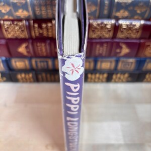 Puede incluir: Un libro titulado "Pippi Longstocking" con una columna vertebral morada. La columna vertebral presenta texto blanco y un dise&ntilde;o floral. Las p&aacute;ginas del libro son visibles, y el fondo muestra una pila de otros libros.