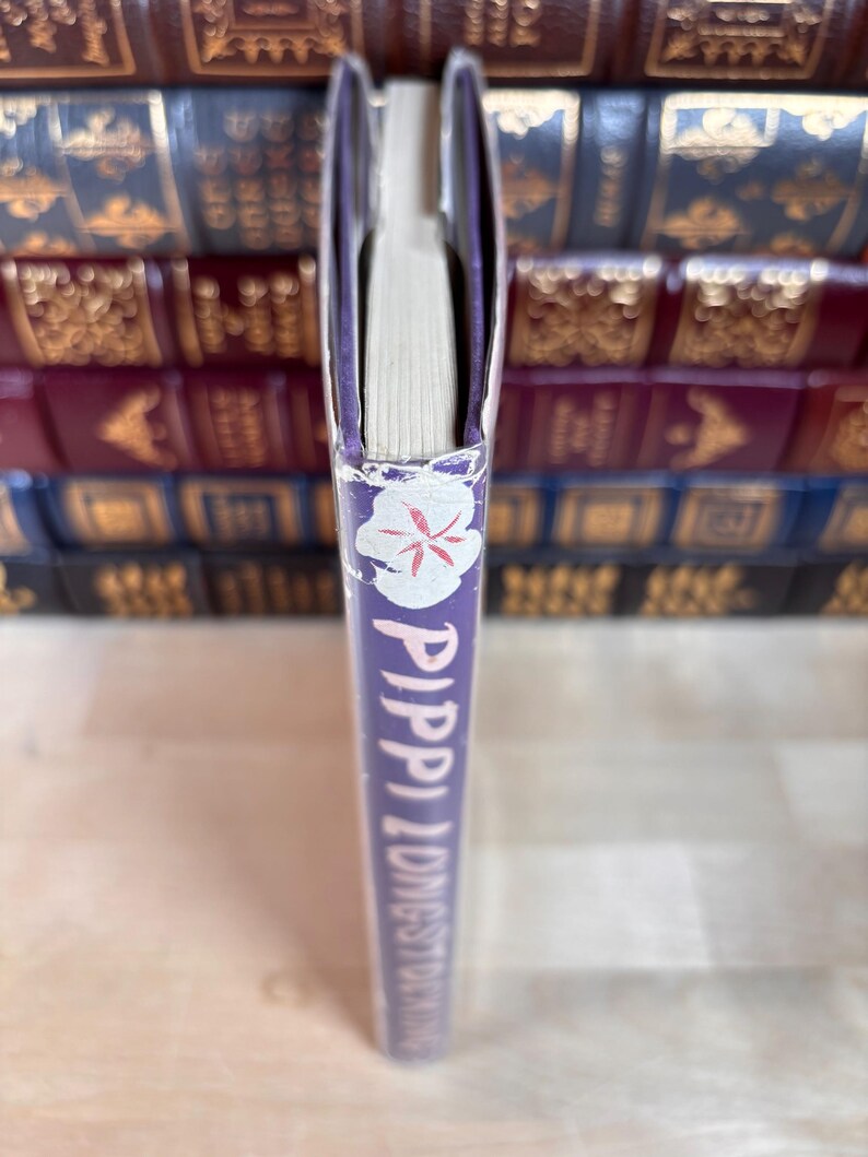 Puede incluir: Un libro titulado "Pippi Longstocking" con una columna vertebral morada. La columna vertebral presenta texto blanco y un dise&ntilde;o floral. Las p&aacute;ginas del libro son visibles, y el fondo muestra una pila de otros libros.