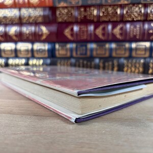 Puede incluir: Un libro de tapa dura con lomo morado y cubierta colorida, apoyado sobre una superficie de madera. Las p&aacute;ginas del libro est&aacute;n envejecidas y se ve un marcap&aacute;ginas. Una pila de otros libros est&aacute; al fondo.