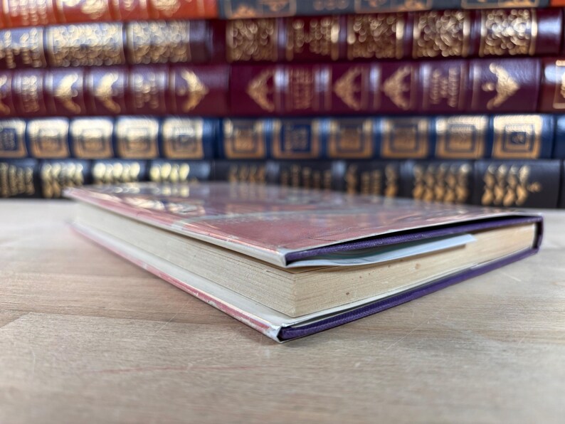 Puede incluir: Un libro de tapa dura con lomo morado y cubierta colorida, apoyado sobre una superficie de madera. Las p&aacute;ginas del libro est&aacute;n envejecidas y se ve un marcap&aacute;ginas. Una pila de otros libros est&aacute; al fondo.