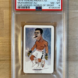 May include: A vintage trading card featuring a cartoon illustration of Muhammad Ali, a famous boxer, with the text "The Greatest" on his boxing shorts. The card is in a protective plastic case with a label that reads "1979 Venorlandus LTD. #3 Muhammad Ali NM-MT Our Heroes World of Sport 67282036".