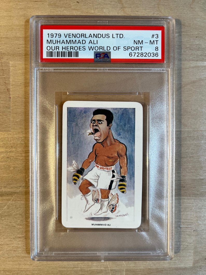 May include: A vintage trading card featuring a cartoon illustration of Muhammad Ali, a famous boxer, with the text "The Greatest" on his boxing shorts. The card is in a protective plastic case with a label that reads "1979 Venorlandus LTD. #3 Muhammad Ali NM-MT Our Heroes World of Sport 67282036".