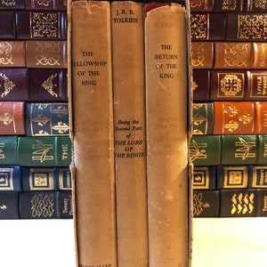 May include: A set of three vintage hardcover books, The Fellowship of the Ring, The Two Towers, and The Return of the King, by J.R.R. Tolkien. The books are bound in brown paper with gold lettering and are part of a larger collection of books.