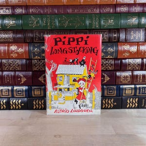 May include: A copy of the book Pippi Longstocking by Astrid Lindgren with a bright red cover featuring a black and white illustration of a house, a ladder, and a character with braided hair. The book is positioned in front of a shelf of leather-bound books.