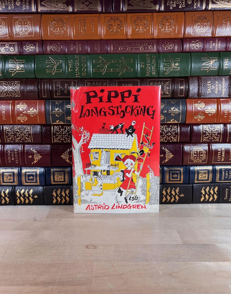 May include: A copy of the book Pippi Longstocking by Astrid Lindgren with a bright red cover featuring a black and white illustration of a house, a ladder, and a character with braided hair. The book is positioned in front of a shelf of leather-bound books.