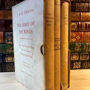 May include: A three-volume set of The Lord of the Rings by J.R.R. Tolkien. The books are bound in brown cloth with gold lettering. The set is in a slipcase with a white background and red lettering.