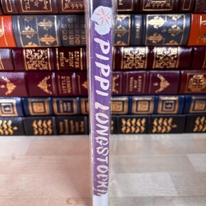 May include: A purple book spine with the title Pippi Longstocking printed in white text, featuring a small white flower graphic at the top. The book is wrapped in clear plastic and positioned vertically in front of a stack of leather-bound books.