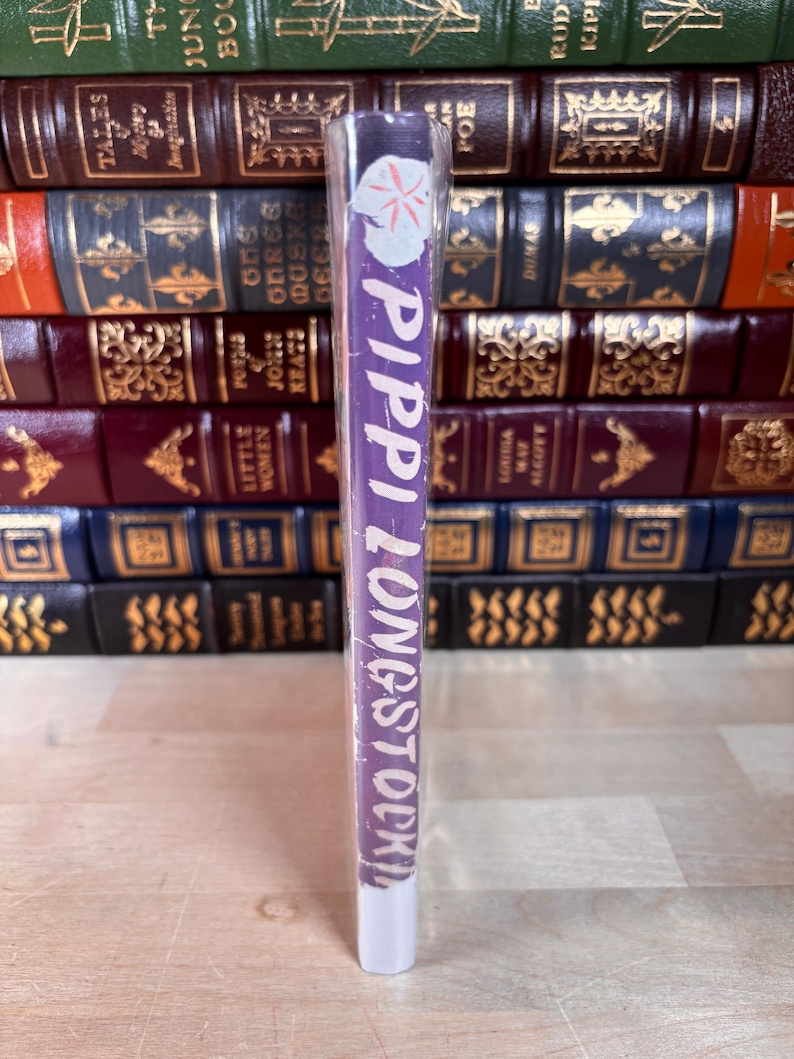 May include: A purple book spine with the title Pippi Longstocking printed in white text, featuring a small white flower graphic at the top. The book is wrapped in clear plastic and positioned vertically in front of a stack of leather-bound books.