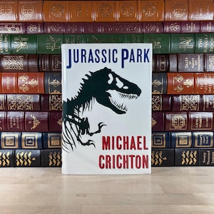 May include: A hardcover copy of "Jurassic Park" by Michael Crichton stands in front of a stack of vintage books. The book cover is white with the title in blue and the author's name in red. A black dinosaur silhouette is featured.