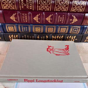 May include: A light gray hardcover book titled Pippi Longstocking by Lindgren with a red line drawing of a character on the front cover, positioned in front of a stack of various leather-bound books with gold embossed spines and lettering.