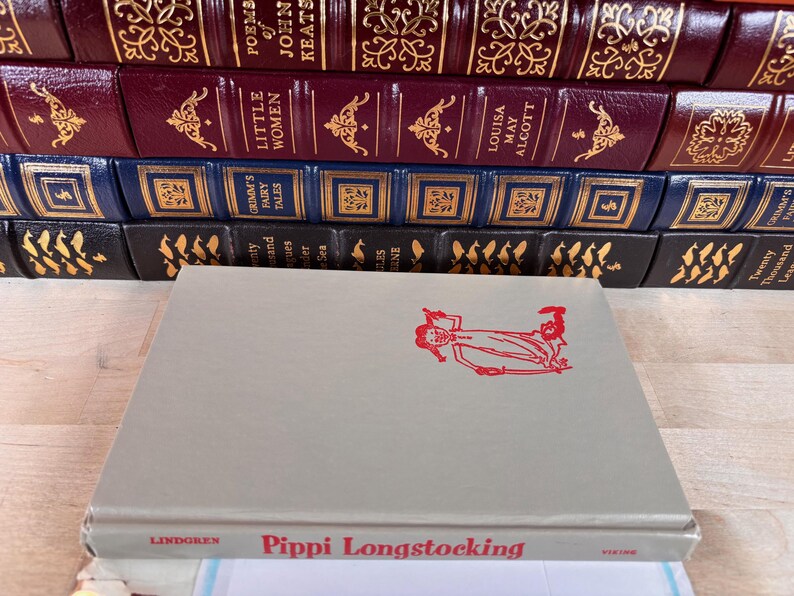 May include: A light gray hardcover book titled Pippi Longstocking by Lindgren with a red line drawing of a character on the front cover, positioned in front of a stack of various leather-bound books with gold embossed spines and lettering.