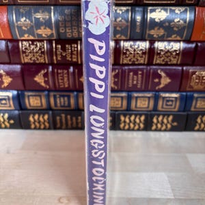 Puede incluir: Un libro de tapa dura titulado "Pippi Longstocking" con lomo morado. El lomo presenta texto blanco y un dise&ntilde;o floral. El libro se muestra frente a otros libros antiguos.
