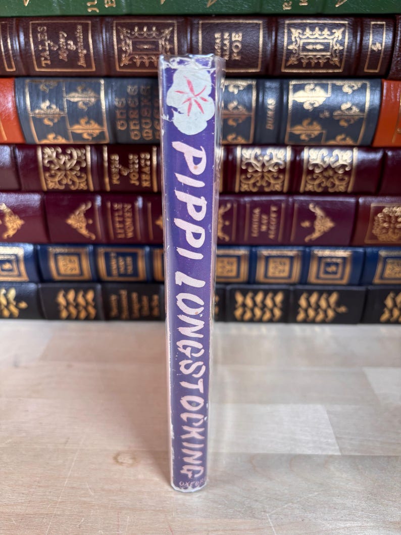 Puede incluir: Un libro de tapa dura titulado "Pippi Longstocking" con lomo morado. El lomo presenta texto blanco y un dise&ntilde;o floral. El libro se muestra frente a otros libros antiguos.
