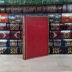 May include: A red hardcover book with gold detailing is positioned in front of a stack of antique books. The book's cover is plain red, and the spine features gold lettering. The other books have diverse colours and designs.
