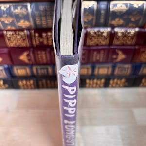 May include: A purple book spine with the title Pippi Longstocking printed in white vertical text. A small white circular icon with a red flower design is positioned at the top of the spine. The book is shown from a side profile view.