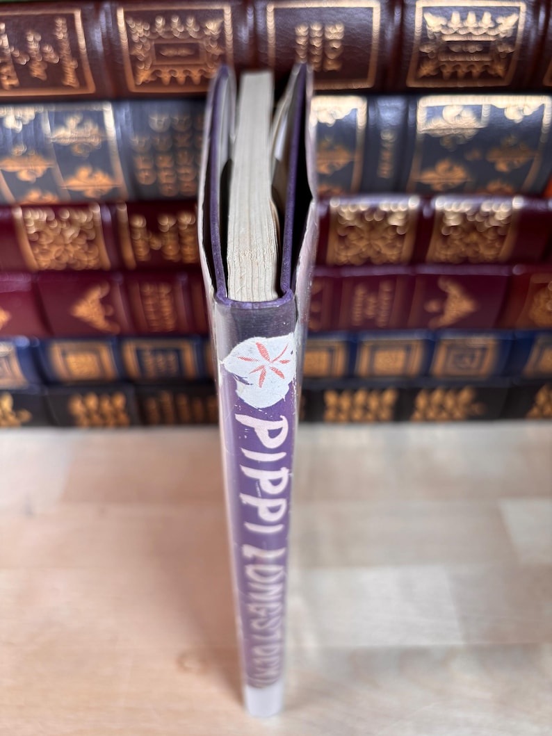 May include: A purple book spine with the title Pippi Longstocking printed in white vertical text. A small white circular icon with a red flower design is positioned at the top of the spine. The book is shown from a side profile view.