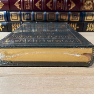 May include: A black hardcover book with gold lettering on the spine. The book is wrapped in plastic and has a gold-colored top edge.