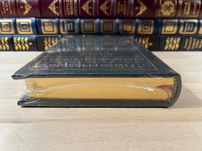 May include: A black hardcover book with gold lettering on the spine. The book is wrapped in plastic and has a gold-colored top edge.