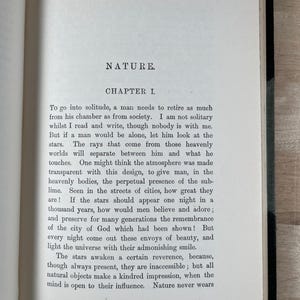 May include: Open book with text titled "NATURE" and "CHAPTER I". The text describes the need for solitude and the beauty of the stars. The pages are aged with a slightly yellowed tone, and the text is in black.
