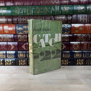 May include: A hardcover book titled "Catch-22" by Joseph Heller, with a green cover and white lettering. The book is standing in front of a collection of antique books with various colors and gold lettering.
