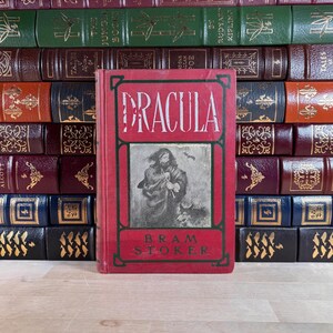 May include: A red hardcover book titled "DRACULA" by Bram Stoker, with an illustration of a man and a dog. The book is in front of a stack of various antique books with leather covers in shades of green, brown, and burgundy.