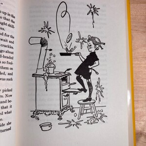 May include: A black and white line drawing illustration on a book page depicting a person standing on a small stool, cooking at a stove. The person holds a frying pan with a pancake flipping in the air, while bubbles and splashes surround the scene.