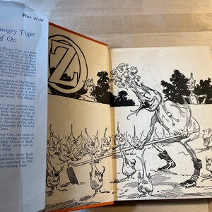 K&ouml;nnte beinhalten: Ein offenes Buch mit einer Schwarzwei&szlig;illustration einer Figur aus dem Buch "Der hungrige Tiger von Oz". Die Figur ist eine humanoide Gestalt mit einem gro&szlig;en Kopf und einem langen, d&uuml;nnen K&ouml;rper. Die Figur h&auml;lt einen langen, d&uuml;nnen Stock in ihrer rechten Hand. Die Figur steht in einer Waldlandschaft. Der Buchumschlag tr&auml;gt den Titel "Der hungrige Tiger von Oz" und den Preis "1,50 &euro;".