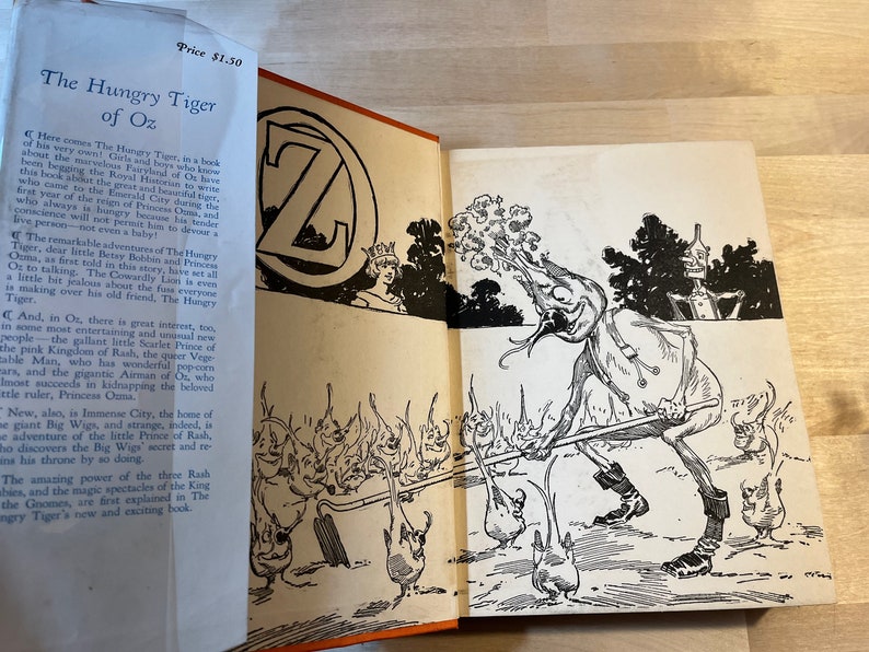 K&ouml;nnte beinhalten: Ein offenes Buch mit einer Schwarzwei&szlig;illustration einer Figur aus dem Buch "Der hungrige Tiger von Oz". Die Figur ist eine humanoide Gestalt mit einem gro&szlig;en Kopf und einem langen, d&uuml;nnen K&ouml;rper. Die Figur h&auml;lt einen langen, d&uuml;nnen Stock in ihrer rechten Hand. Die Figur steht in einer Waldlandschaft. Der Buchumschlag tr&auml;gt den Titel "Der hungrige Tiger von Oz" und den Preis "1,50 &euro;".