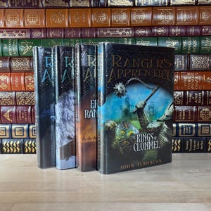 May include: A stack of hardcover books, with a focus on a series titled "Ranger's Apprentice." The covers feature illustrations of fantasy scenes. The book "The Kings of Clonmel" by John Flanagan is visible. The background includes a collection of antique books.