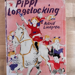 May include: A hardcover book titled Pippi Longstocking by Astrid Lindgren with a pink cover illustration featuring a girl riding a white horse, a monkey on her shoulder, and several children running alongside in a colorful, illustrated scene.
