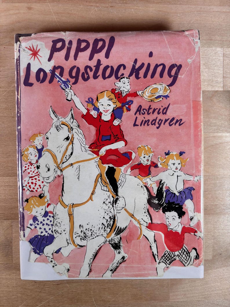 May include: A hardcover book titled Pippi Longstocking by Astrid Lindgren with a pink cover illustration featuring a girl riding a white horse, a monkey on her shoulder, and several children running alongside in a colorful, illustrated scene.
