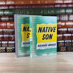 Può includere: È esposto un esemplare di "Native Son" di Richard Wright. Il libro ha una copertina gialla e blu con il titolo in grandi lettere. È presentato davanti a una collezione di altri libri d'epoca.
