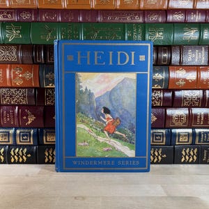 Op de afbeelding: Een blauw hardcover boek met de titel "HEIDI" met gouden letters, onderdeel van de "WINDERMERE SERIES". De omslag toont een kleurrijke illustratie van een meisje dat door een berglandschap rent. De achtergrond bestaat uit een stapel vintage boeken.