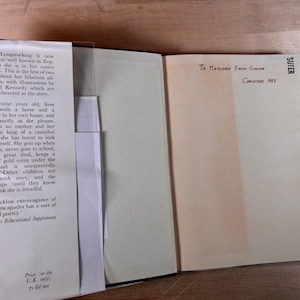 May include: An open hardcover book with a cream-colored dust jacket flap on the left featuring black printed text about Pippi Longstocking. The right page is blank with handwritten ink text reading To Marjorie From Gunvor Christmas 1957.