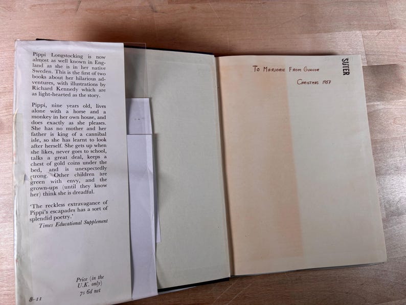 May include: An open hardcover book with a cream-colored dust jacket flap on the left featuring black printed text about Pippi Longstocking. The right page is blank with handwritten ink text reading To Marjorie From Gunvor Christmas 1957.